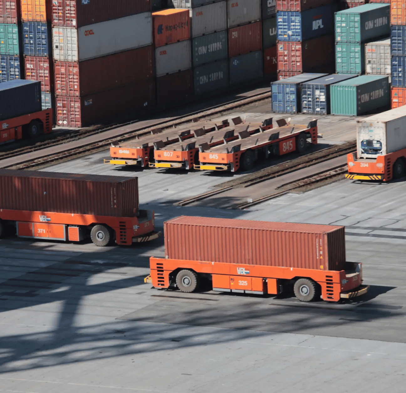 Shipping containers being hauled around a shipyard, many containers stacked in background. 